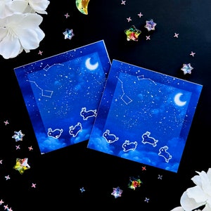 Op de afbeelding: Twee blauwe kaarten met een ontwerp van een sterrenhemel en silhouetten van springende konijnen. De kaarten tonen een halve maan en een sterrenbeeld aan de hemel.