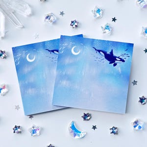 May include: Two square sticky notes featuring a blue gradient background with a crescent moon and an orca whale swimming among small fish. The design includes light streaks and a soft, ethereal atmosphere across the surface of the paper sheets.