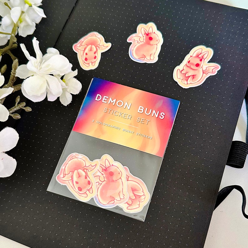May include: A set of three holographic bunny stickers with demon horns and tails. The stickers are packaged in a clear plastic bag with a colorful label that reads "Demon Buns Sticker Set 3 Holographic Bunny Stickers".