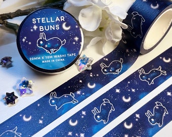 May include: A roll of washi tape with a blue starry night sky design featuring white bunnies and crescent moons. The tape is 20mm wide and 10m long. The label on the tape reads "Stellar Buns 20mm x 10m Washi Tape Made in China."