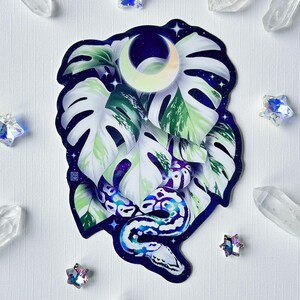 May include: A sticker featuring a white and green monstera leaf with a blue and white snake curled around it. The background is a dark blue with white stars and a crescent moon.