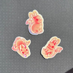 May include: Three holographic stickers featuring a pink cartoon bunny with wings. The bunny is in different poses on each sticker.