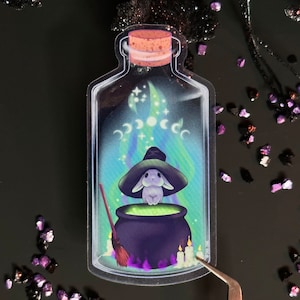 May include: A clear glass bottle sticker with a white rabbit wearing a witch's hat and holding a cauldron with green liquid. The bottle is filled with purple and blue smoke and has a cork stopper. There are three crescent moons and stars in the smoke.