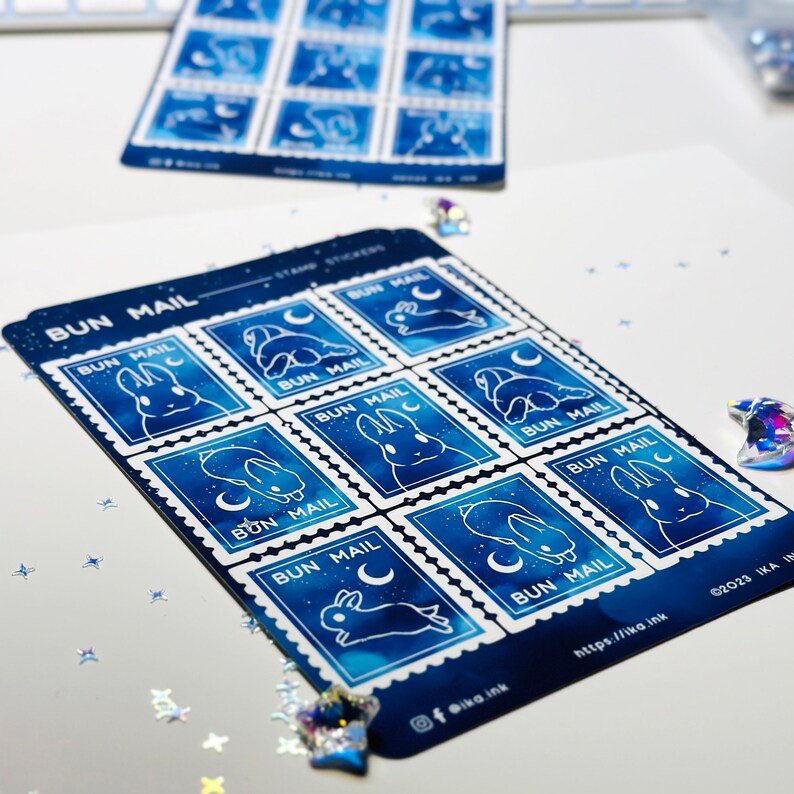 May include: A sheet of eight blue and white bunny stamp stickers with the text "BUN MAIL" on each stamp. The stamps feature a white bunny with a crescent moon and stars on a blue watercolour background.
