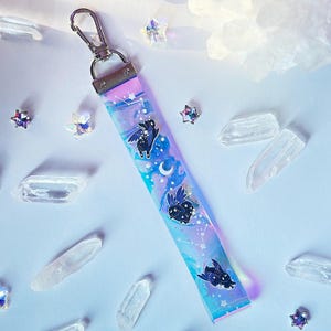 May include: A celestial-themed keychain wristlet with a silver clasp. The wristlet features a gradient of pink, blue, and purple with a constellation design and black winged bunny illustrations. Clear quartz crystals and star-shaped embellishments surround the wristlet.