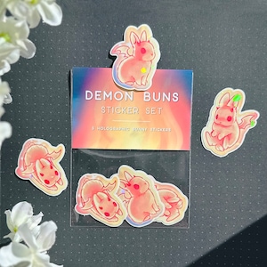 May include: A set of five holographic stickers featuring pink bunny rabbits with wings. The stickers are packaged in a clear plastic bag with a colorful background and the text "Demon Buns Sticker Set 3 Holographic Bunny Stickers".