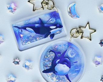 Könnte beinhalten: Zwei Schlüsselanhänger aus Acryl mit Orca-Wal-Designs. Einer ist rechteckig, der andere rund. Beide haben ein blau-weißes Farbschema mit glitzerndem Hintergrund. Goldene sternförmige Schlüsselringe und dekorative Kristalle umgeben die Schlüsselanhänger.