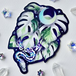 Pu&ograve; includere: Un adesivo con una foglia di monstera bianca e verde con un serpente blu e viola avvolto attorno. Lo sfondo &egrave; blu scuro con stelle bianche e una luna crescente.