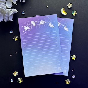 May include: Two sheets of lined paper with a gradient background of purple and blue. The paper has a white outline of bunnies hopping in circles with stars scattered across the background. The paper has the text "Tako Ink" in a small square at the bottom right corner.