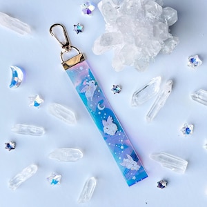 May include: A blue and purple iridescent keychain with a gold clasp. The keychain features a design of three white bunnies with wings flying against a background of stars and a crescent moon.