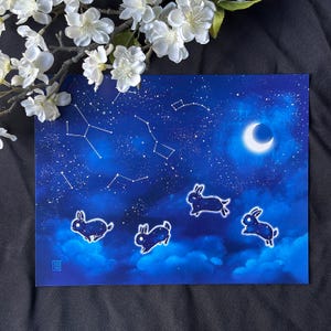 以下が含まれることがあります： 星と三日月のある夜空の青い水彩画。4匹の黒いウサギのシルエットが空を跳ねています。
