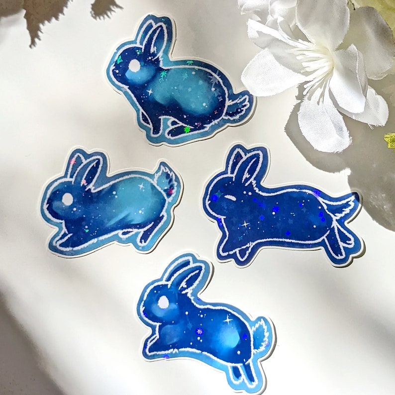 May include: Four stickers featuring a blue and white galaxy bunny design. The stickers have a holographic effect, with the bunnies in various running poses. The stickers are on a white background with a white flower.