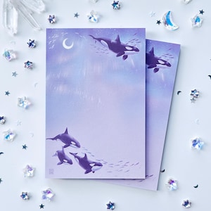 May include: Two rectangular sheets of stationery featuring a gradient purple and blue background with illustrations of orcas swimming. The design includes a crescent moon, small fish, and a subtle light effect. Each sheet has a small square logo.