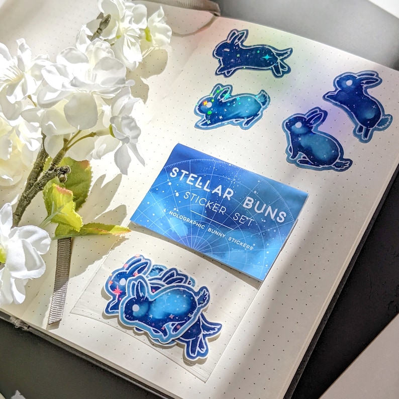 May include: A set of holographic bunny stickers with a celestial design, featuring shades of blue and sparkling accents. The stickers are displayed on a dotted notebook next to white flowers. The sticker set packaging reads "Stellar Buns".