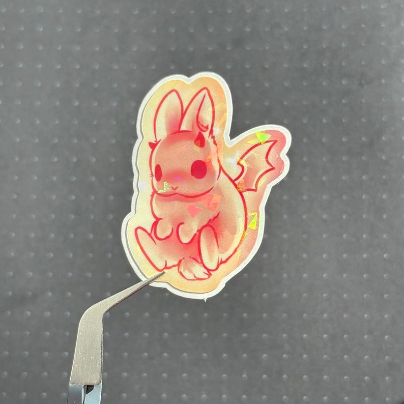 May include: A pink and white iridescent sticker of a cartoon bunny with devil horns and wings. The bunny is sitting with its legs crossed and has a cute expression on its face.