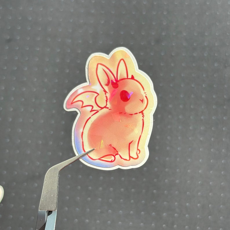 May include: A holographic sticker featuring a pink and orange cartoon bunny with wings and a devil tail. The bunny has red eyes and is sitting on its hind legs.