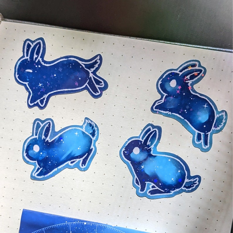 May include: Four blue bunny stickers with a galaxy design. Each sticker features a different pose of a rabbit with a dark blue body filled with stars and a white outline. The stickers are on a light gray surface.