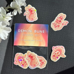 May include: A clear plastic bag with a holographic sticker set featuring 5 pink and white demon bunny stickers. The bag has a label that reads "Demon Buns Sticker Set" and "3 Holographic Bunny Stickers".