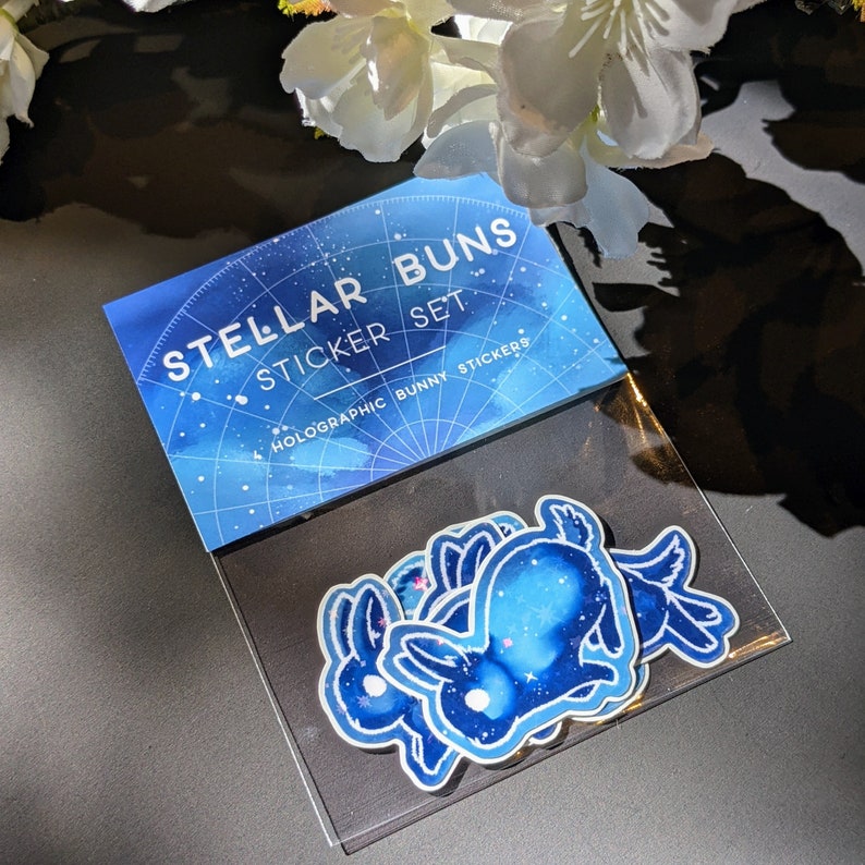 May include: A set of holographic bunny stickers in a clear plastic sleeve. The stickers feature a blue and white galaxy design. The packaging reads "Stellar Buns Sticker Set".