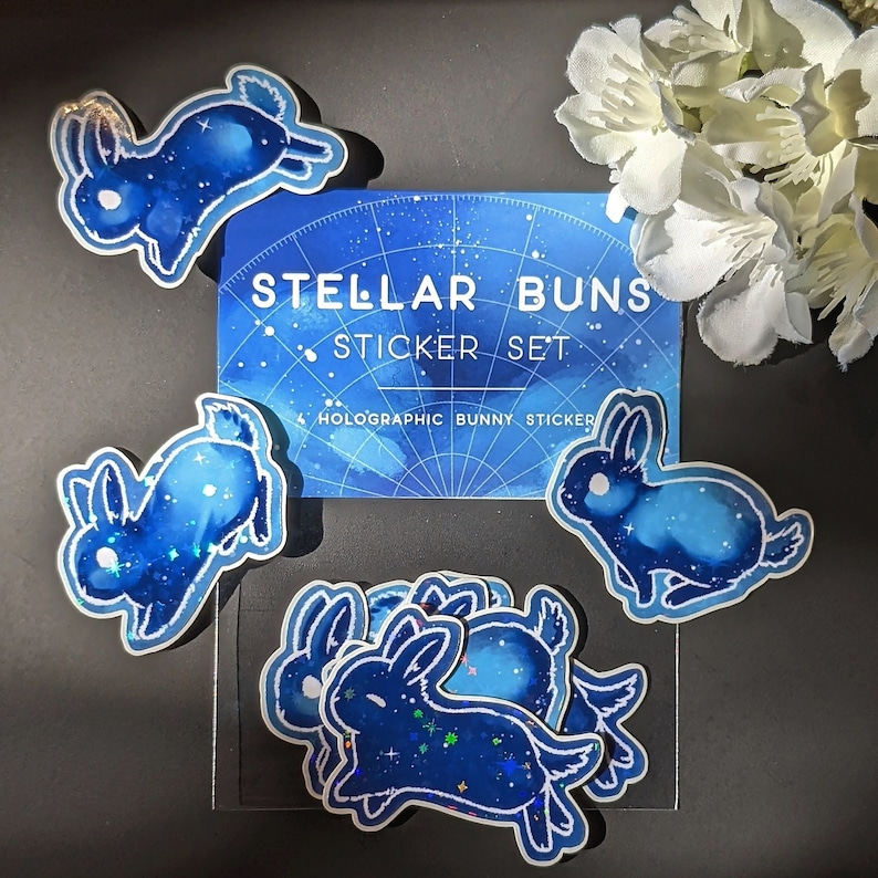 May include: A set of holographic bunny stickers with a celestial design. The stickers feature blue watercolor bunnies with a starry night sky pattern. The set includes a card that reads "Stellar Buns Sticker Set".