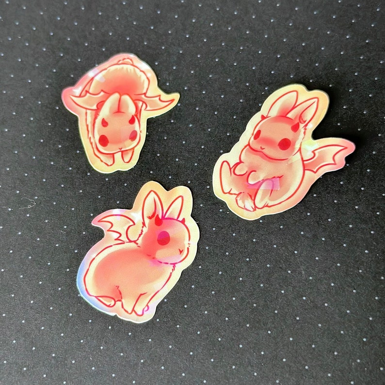 May include: Three iridescent stickers featuring pink and white cartoon bunnies with wings and horns. The bunnies are in different poses, one is sitting, one is standing, and one is upside down.
