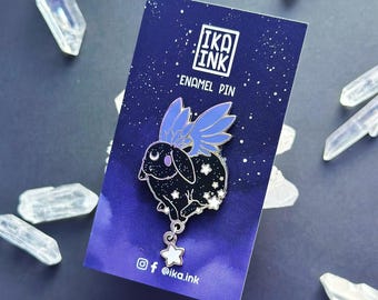 May include: A black enamel pin of a rabbit with blue wings and a star charm. The rabbit is covered in white stars and is set against a dark blue starry sky background. The pin says "IKA INK Enamel Pin" and "@ika.ink" on the bottom.