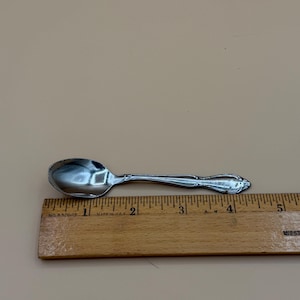 May include: A small, shiny silver spoon with a decorative handle, placed next to a wooden ruler. The spoon is approximately 4 inches long. The ruler is marked with inch measurements.