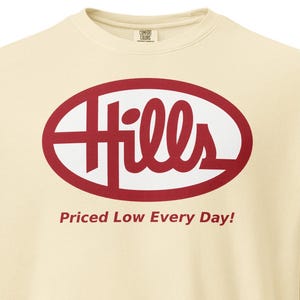 Op de afbeelding: Crèmekleurige sweatshirt met lange mouwen. De voorkant heeft een rood ovaal logo met het woord "Hills" in wit en de tekst "Priced Low Every Day!" eronder. De sweatshirt heeft een ronde hals en een relaxte pasvorm.