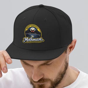 Puede incluir: Gorra de béisbol negra con visera plana. La gorra presenta un parche circular con las palabras "West Virginia" y "Mothman" en una fuente estilizada. El parche incluye una ilustración de una criatura de ojos rojos.