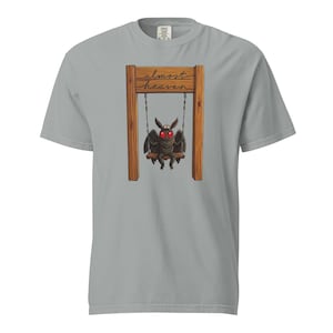 May include: A light gray t-shirt featuring a cartoon moth on a swing set. The moth has red eyes and is sitting on a wooden swing. The words "almost heaven" are written above the moth in a cursive font.