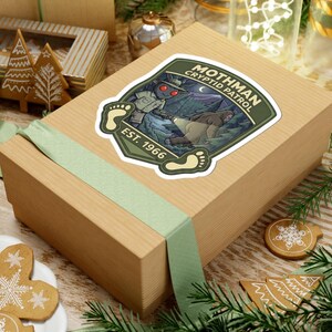 May include: A sticker featuring the Mothman Cryptid Patrol logo, with a green and white design. The sticker is on a brown gift box with a green ribbon. The logo includes the text "Mothman Cryptid Patrol" and "EST. 1966".