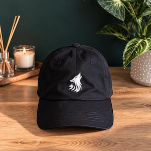 May include: A black baseball cap featuring a white embroidered graphic of a howling wolf on the front center panel. The cap has a structured crown, a curved brim, and a button at the top. The design is centered and stitched in white thread.