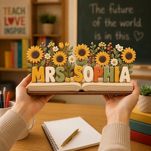May include: A decorative wooden display featuring an open book base with the text MRS. SOPHIA in raised, multi-colored block letters. Behind the text, a row of sculpted sunflowers, small white daisies, and green foliage creates a floral arrangement.