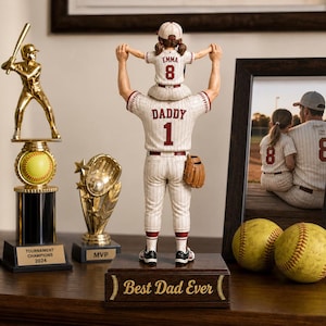 Op de afbeelding: Een honkbalthema-opstelling met een trofee, een beeldje van een honkbalspeler met een kind en ingelijste foto's. Het beeldje heeft de tekst "DADDY 1" op de rug. Er zijn ook twee honkballen en een houten bord met "Best Dad Ever".