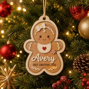 May include: A layered wooden gingerbread person ornament featuring a white bow, a bib with a red heart, and a small peppermint candy. The text reads Avery and FIRST CHRISTMAS 2026. The ornament is suspended by a tan string against a pine tree background.