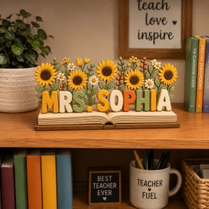 May include: A decorative wooden display featuring an open book base with the text MRS. SOPHIA in raised, colorful letters. The display is adorned with sculpted sunflowers, daisies, and green foliage, creating a floral arrangement above the name.