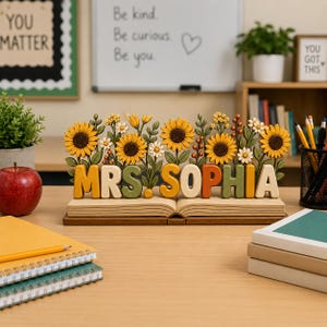 May include: A wooden desk sign featuring the name MRS. SOPHIA in raised, multi-colored block letters. The sign is shaped like an open book and is decorated with carved sunflowers, small white flowers, and green foliage along the top edge.