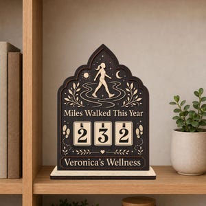 Op de afbeelding: Een donkerbruine houten tracker met een spitsboogvormige bovenkant, voorzien van een silhouet van een lopend persoon, de tekst "Miles Walked This Year", drie draaibare cijfertegels met "232" en onderaan "Veronica's Wellness".