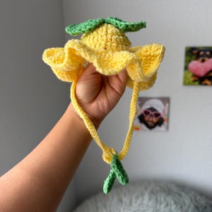 May include: A handmade crochet hat in a bright yellow color, resembling a flower. The hat features a ruffled brim and green leaf accents on top and at the chin ties. The hat is held up by a hand.