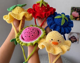 May include: Five crocheted hats featuring fruit and animal designs including a yellow lemon, a red strawberry, a blue blueberry, a pink watermelon slice with green rind, and a yellow duck with an orange beak, all with chin straps.