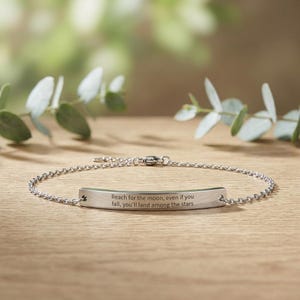 May include: A silver bracelet featuring a chain and a rectangular bar. The bar is engraved with the words "Reach for the moon, even if you fall, you'll land among the stars." The bracelet is displayed on a wooden surface with green foliage.