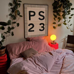 May include: A bedroom scene featuring a bed with pink bedding and striped pillows. A large wall hanging with the letters "PS 23" is above the bed. A small lamp with a yellow globe sits on a red stand.