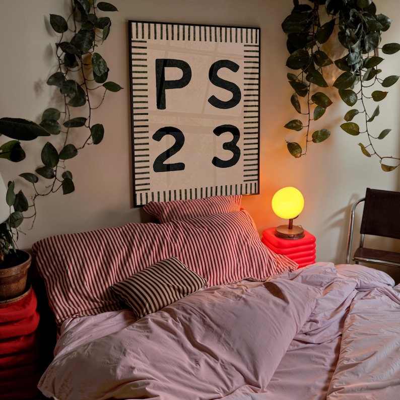 May include: A bedroom scene featuring a bed with pink bedding and striped pillows. A large wall hanging with the letters "PS 23" is above the bed. A small lamp with a yellow globe sits on a red stand.