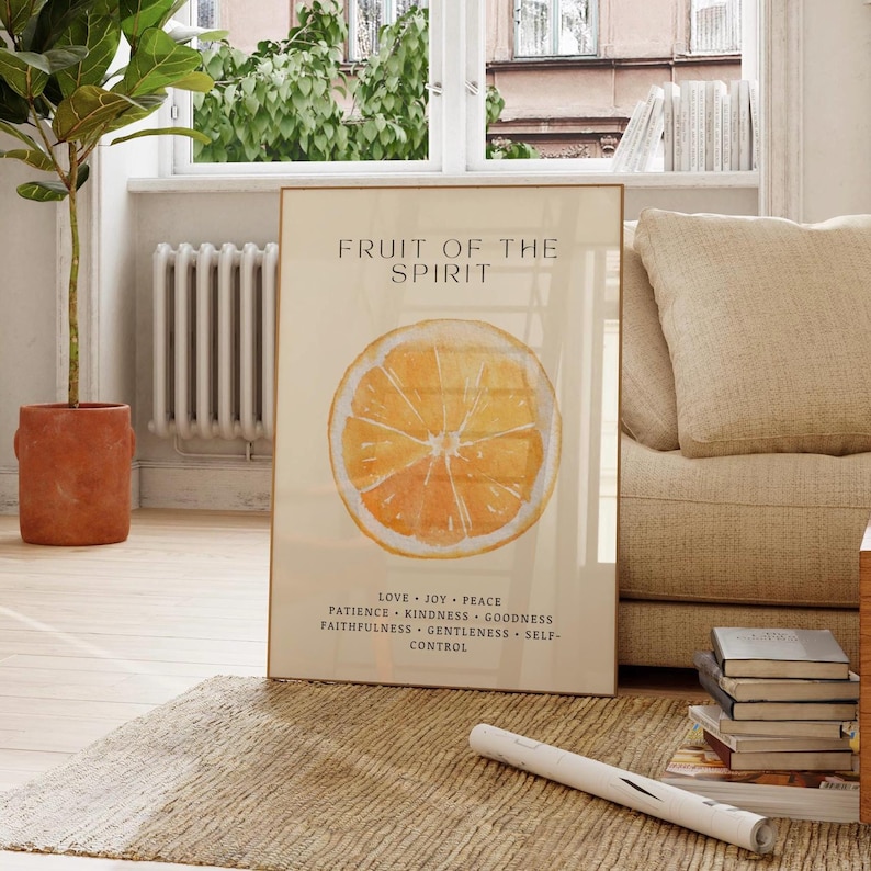 A framed “Fruit of the Spirit” poster featuring a bright watercolor orange rests on a beige sofa in a sunlit living room. Soft natural light enhances the warm citrus tones and green leaf details. A woven rug, wooden floor, and stacked books create a cozy, inviting setting that highlights the Christian virtues listed below the artwork.