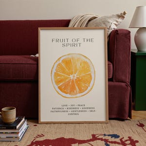 A framed “Fruit of the Spirit” orange poster stands near a deep red couch, with a folded blanket and stacked books adding warmth to the room. Soft natural light illuminates the watercolor orange slice and leafy accents. The Christian virtues at the bottom create a calming, encouraging presence in the cozy space.