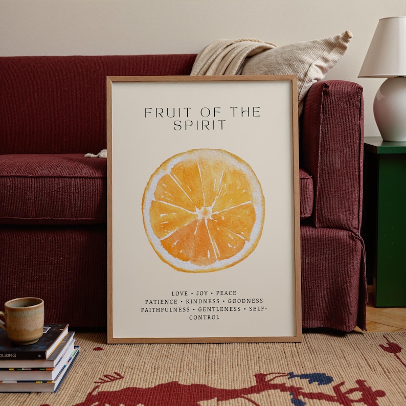 A framed “Fruit of the Spirit” orange poster stands near a deep red couch, with a folded blanket and stacked books adding warmth to the room. Soft natural light illuminates the watercolor orange slice and leafy accents. The Christian virtues at the bottom create a calming, encouraging presence in the cozy space.