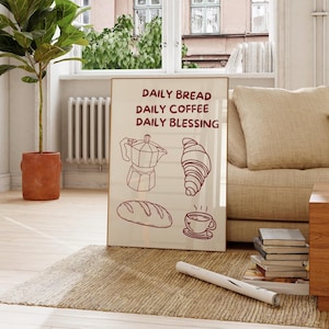 Könnte beinhalten: Ein gerahmter Druck mit den Worten "DAILY BREAD, DAILY COFFEE, DAILY BLESSING" in braunem Text. Der Druck zeigt Strichzeichnungen einer Kaffeemaschine, eines Croissants, eines Brotlaibs und einer Tasse Kaffee. Der Rahmen ist hellbraun.