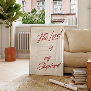 May include: A framed print with the text "The Lord is my Shepherd" in red script. The print is displayed on a light-colored wall, with a beige sofa and a potted plant in the background. The room has a neutral color palette.