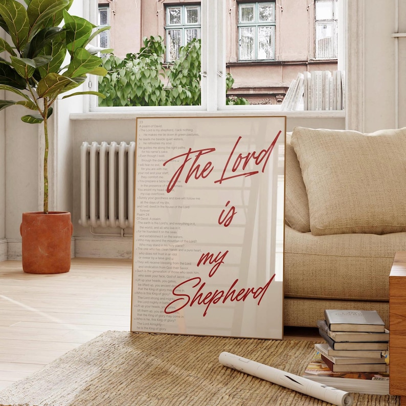 May include: A framed print with the text "The Lord is my Shepherd" in red script. The print is displayed on a light-colored wall, with a beige sofa and a potted plant in the background. The room has a neutral color palette.