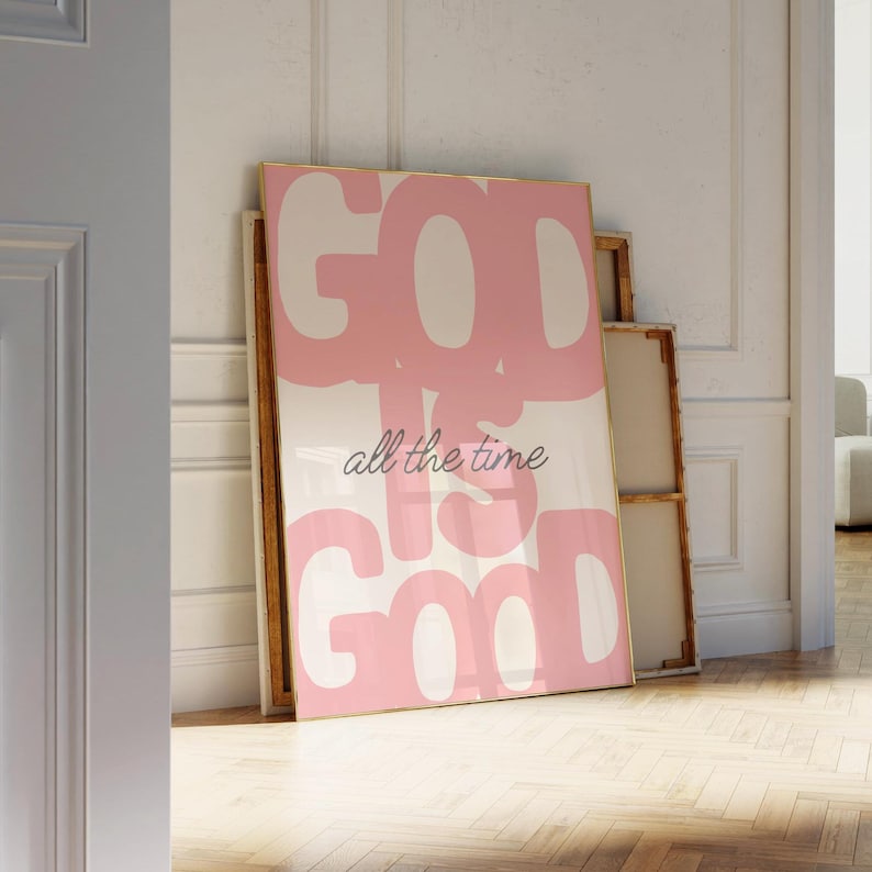 May include: A framed art print with the words "GOD IS GOOD all the time" in a pink and white design. The artwork is leaning against a wall, with a gold frame and a minimalist aesthetic.